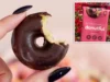 Better Bakehouse 正在召回因过敏反应而贴错标签的巧克力甜甜圈 A hand with dark fingernails holds a chocolate-dipped vanilla cake donut with a bite taken out of it.